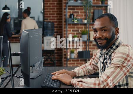 Database programmer writing code and smiling while coworker walks by holding clipboard in it agency, relaxed app developer typing on keyboard with colleagues computing big data. Stock Photo