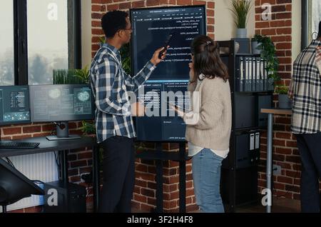 Database administrators compare source code on a wall screen and tablet in a busy server room while cloud programmers debug software algorithms. Stock Photo