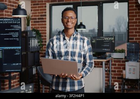African American programmer stands with a laptop and smiles toward the camera while coworkers compute big data in an IT agency office. Stock Photo