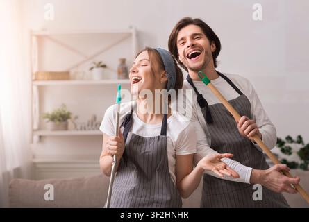 Cheerful Couple Using Mop Handles As Microphones, Having Fun During Spring-Cleaning Stock Photo