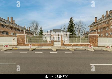 Asphalt road with parking bollards in front of a brick wall and white fence with a central gatehouse, flanked by large brick apartment buildings. Stock Photo