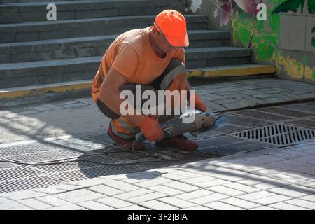 Kyiv, Ukraine - July 17, 2026: A road maintenance worker uses a handheld angle grinder to cut a metal storm drain grate during street repair work at V Stock Photo