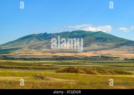Vast open fields stretch toward rolling hills under clear blue sky. Gentle slopes rise in distance, covered in green vegetation Stock Photo