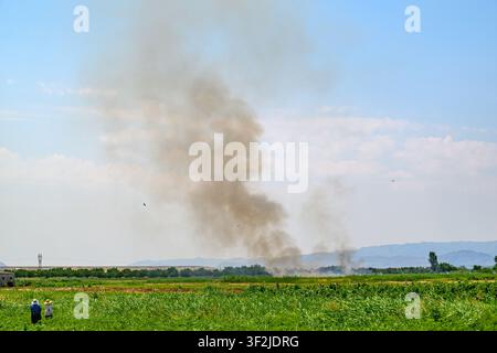 Large plume of smoke rises over green field. Two people walk near edge of field. Sky is partly cloudy. Distant hills visible on horizon Stock Photo