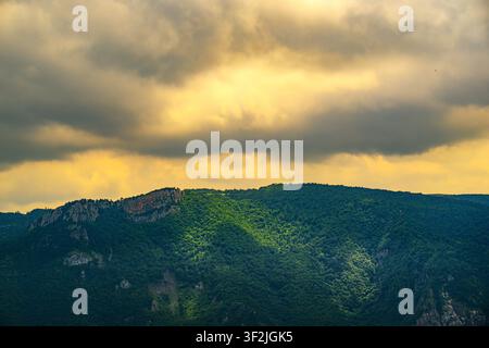 Dramatic sky with golden light breaking through dark clouds. Lush green forest covers rolling hillsides. Mountain ridges rise above dense vegetation Stock Photo