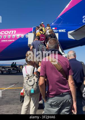 Passengers queueing and walking up stairs to board a pink and blue Wizzair aircraft on a sunny day. Stock Photo