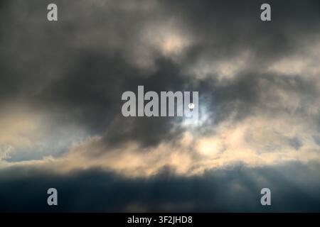 Moon visible through dark, stormy clouds. Clouds illuminated from behind by sunlight. Dramatic sky with soft golden light breaking through Stock Photo