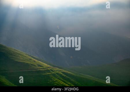 Sunlight breaks through mist over rolling green hills. Shadows stretch across grassy slopes. Valley shrouded in soft clouds Stock Photo