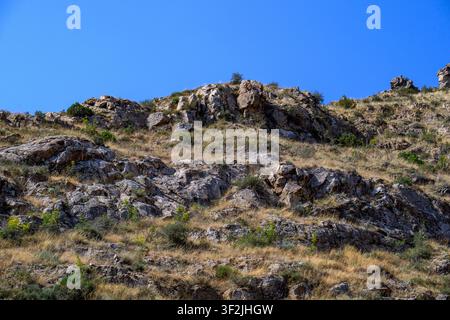 Rugged rocky hillside under clear blue sky. Dry grass and sparse green shrubs cover slope. Sunlight highlights textured stone surfaces Stock Photo