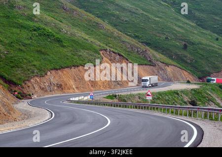 Curved mountain road winds through green hills. Truck drives along asphalt curve. Earthwork exposed on hillside. Guardrails line road edge Stock Photo
