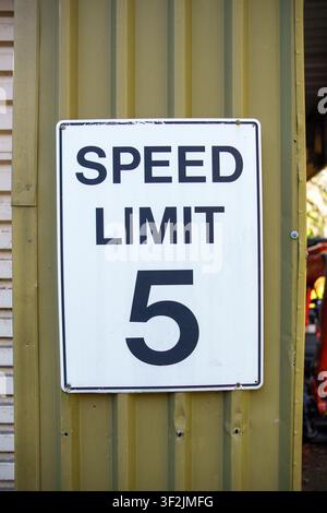 A minimalist white speed limit sign featuring a black five, mounted securely on a weathered olive corrugated metal exterior wall surface. Stock Photo