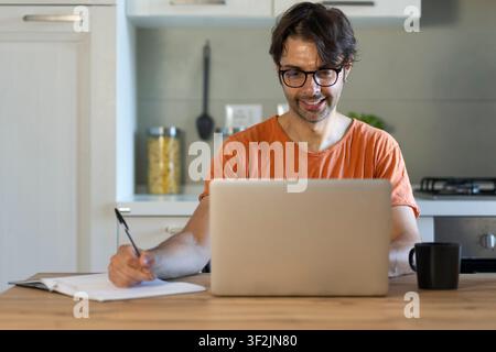 Remote work. Cheerful freelancer guy working on a laptop at home, Sitting at the desk with laptop, making notes, Enjoying freelance Job. Stock Photo