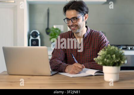 Happy man entrepreneur wearing eyeglasses looking at paper bill checking financial invoice or tax document making online payments working on laptop at Stock Photo
