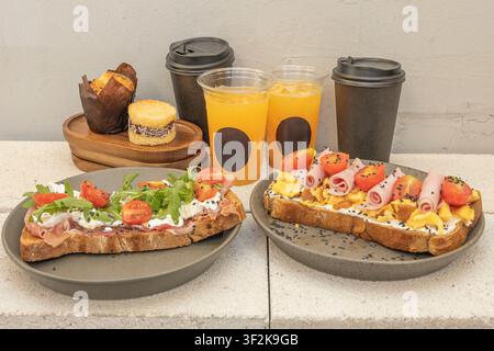 Two savory toasts topped with eggs, ham, tomatoes, and arugula served with orange juice, coffee, and pastries on a gray concrete surface. Stock Photo