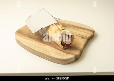 Grilled meat wrap in newspaper-lined paper, filled with steak, tomato, and greens, resting on light wooden serving board. Stock Photo