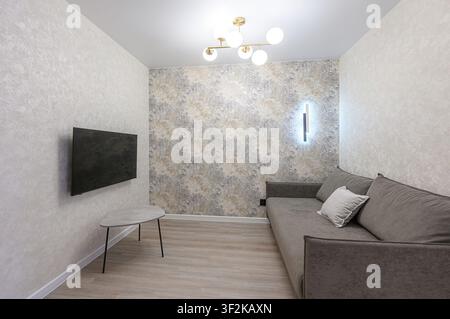 modern living room features a gray sofa, a TV mounted on the wall, and a stylish ceiling light. The room has neutral-toned walls and light-colored flo Stock Photo