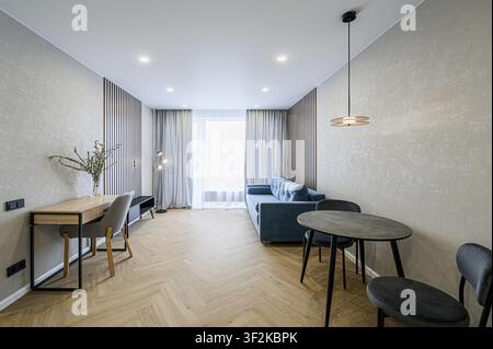 Cozy apartment living room with a blue sofa, dining table, desk, and herringbone floor. Modern design, bright lighting Stock Photo