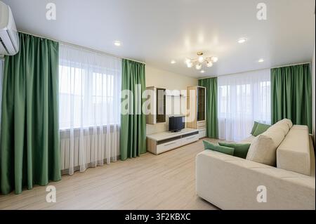 Bright living room with beige sofa, green curtains, and modern furniture. Windows with sheer white curtains let in light Stock Photo