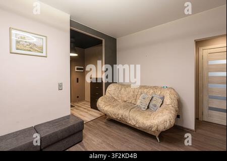 cozy living room featuring a patterned sofa with pillows, gray ottoman, framed picture, wooden floors, and a doorway leading to another room with a dr Stock Photo