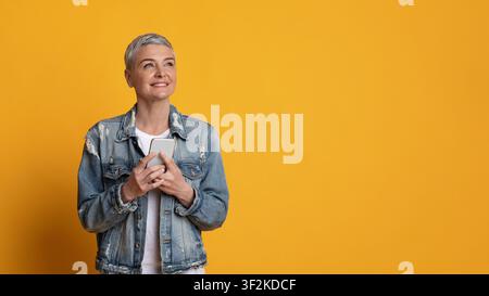 Portrait Of Thoughtful Middle Aged Woman With Smartphone Over Yellow Background Stock Photo