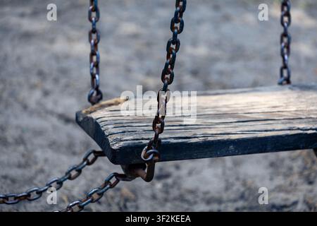 Wooden swing seat hangs from rusted metal chains on a playground frame. The worn surface shows cracks and aged texture. Stock Photo