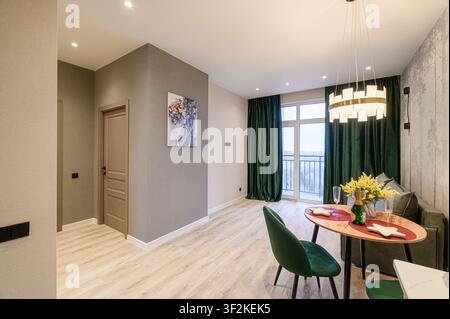 Cozy apartment interior featuring a dining area with an oval table, velvet chairs, and green curtains. The room has modern lighting, light wood floors Stock Photo