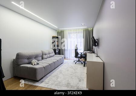 modern, brightly lit room features a gray couch, a desk with a gaming setup, and minimalist décor. The room exudes a functional yet comfortable ambian Stock Photo