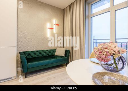 Cozy interior with a green sofa, patterned wallpaper, modern lighting, and a round table adorned with a floral arrangement near a window Stock Photo