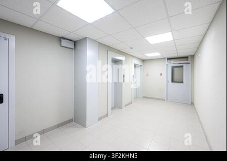 bright corridor with two elevators, tiled floor, and a door. The walls are off-white, and the ceiling has square light panels Stock Photo
