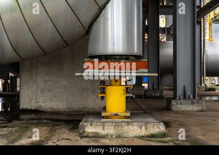 Close up of a yellow pipe support pedestal with an orange anti-vibration pad installed beneath a large industrial storage tank, mounted on a concrete Stock Photo