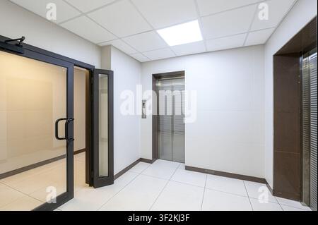 clean, modern interior space featuring white tiled walls and floor. An elevator and an open glass door are visible. The room is well-lit, giving it a Stock Photo