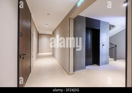 interior shot of a hallway featuring apartment doors, an elevator, and a staircase. The color palette is neutral with beige walls and tiled floors Stock Photo