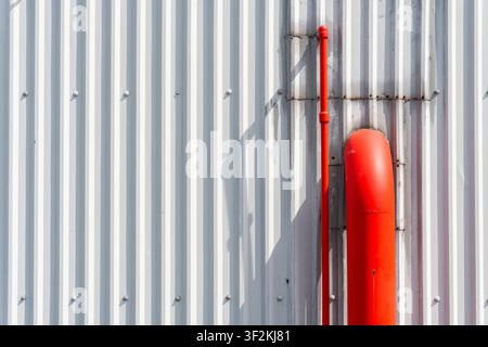 Vibrant red fire suppression pipe mounted on a white corrugated metal wall, creating a bold color contrast with geometric shadows in a minimalist indu Stock Photo