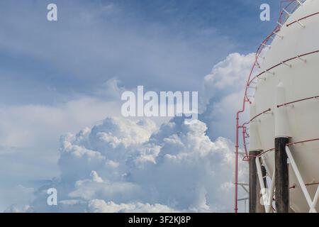 Side view of a large spherical LPG gas storage tank with red pipelines and stairway against a dramatic blue sky with towering cumulus clouds, capturin Stock Photo