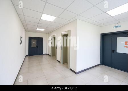 brightly lit hallway features neutral walls, tiled floors, and a suspended ceiling. Two elevators stand between doors. The number "5" is on a wall Stock Photo