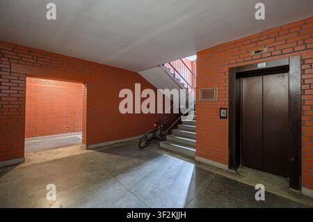 Interior shot of a building hallway with brick walls, a concrete floor, stairs with a bicycle, and an elevator with a display showing the current floo Stock Photo