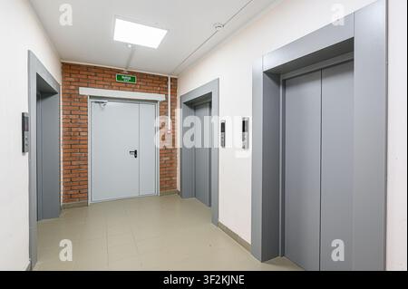 An interior hallway with two elevators, an exit door framed by brick, and tile flooring Stock Photo