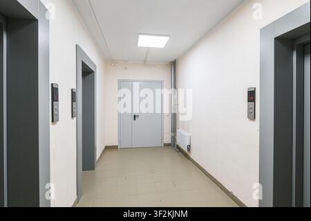 hallway with two elevator doors, a closed metal door, and a tiled floor. The walls are light-colored, and a square light fixture is on the ceiling Stock Photo
