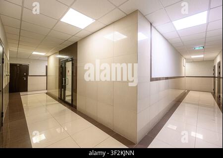 Bright hallway with tiled walls and floors, leading away in two directions. Ceiling lights illuminate the space, and doors line the walls. Exit sign v Stock Photo