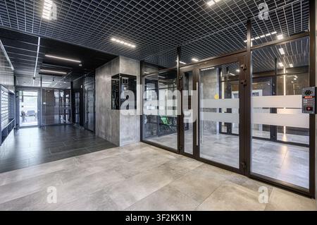 Modern lobby featuring tile floors, glass doors, a grid ceiling with lights, and a security system Stock Photo