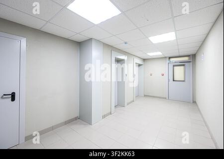 brightly lit hallway with white walls, tiled floor, and a suspended ceiling. There are two elevators and a door visible Stock Photo