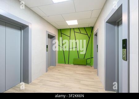 modern hallway with three grey elevators, a green accent wall with black geometric design, light wood-look flooring, and a drop ceiling with fluoresce Stock Photo