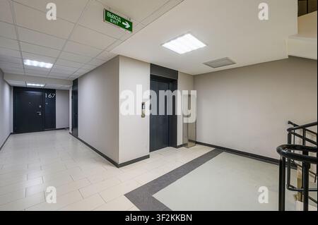 interior shot of a hallway with an elevator, doors, and staircase, featuring tile flooring and neutral-colored walls. Exit sign is visible on the ceil Stock Photo