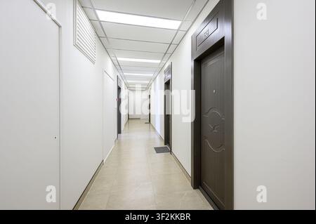 long, well-lit corridor with tiled floor and white walls. Several dark wood doors are visible. Modern ceiling with fluorescent lighting Stock Photo
