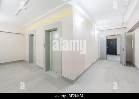 interior shot of a hallway with two elevators on the left and an open doorway to another room on the right. Neutral colors and tile flooring give a cl Stock Photo