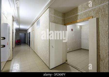 brightly lit hallway in a building. There are doors along the walls, tiled floors, and textured walls. An open doorway reveals an empty room with tile Stock Photo