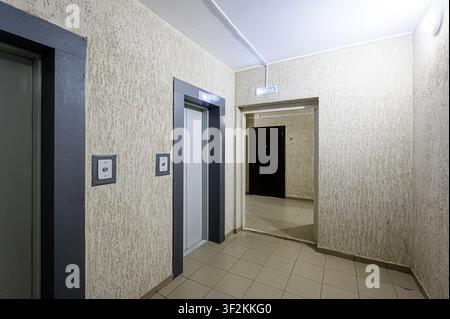 Interior shot of a hallway with beige walls, a tiled floor, and doorways. Two elevators are on the left, and a doorway leads to another room with a bl Stock Photo