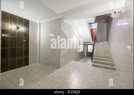 well-lit foyer with tile flooring and walls, leading to stairs. Brown and beige tiles create contrast. A security camera is visible Stock Photo