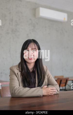 A vertical portrait of a young Asian woman looking directly at the camera with a warm, confident smile. She is wearing a professional beige blazer ove Stock Photo