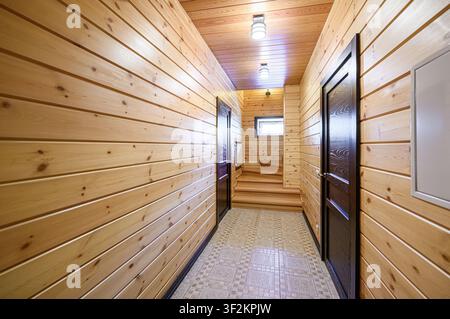 hallway with wooden walls and ceiling, illuminated by ceiling lights. Two dark wooden doors face each other on either side. Stairs are visible at the Stock Photo